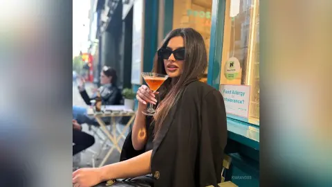 Stylish woman in a black blazer enjoying a cocktail at an outdoor urban cafe on a sunny day.