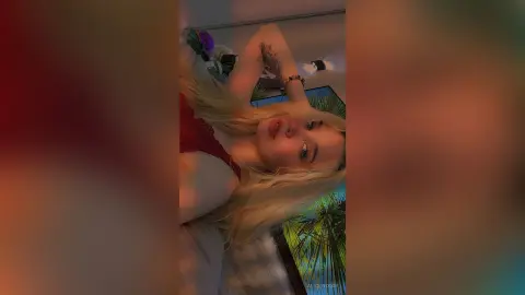 Blonde woman in a red lace top relaxing in a sunlit room with tropical plants and a dreamy mood.