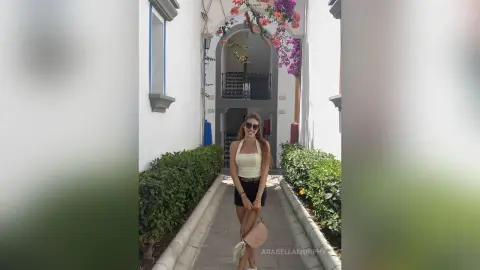 Stylish woman in a white tank top and black shorts walking through a sunny Mediterranean courtyard.