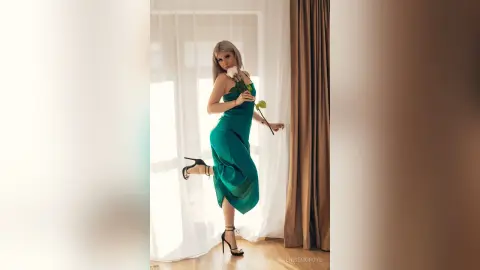 Elegant woman in a green satin dress holding a white rose in a bright, sunlit room.