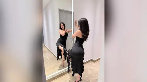Elegant woman in a sleek black midi dress posing in a modern minimalist room mirror reflection.