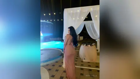 NicoleBella stands in a light pink dress next to a lit swimming pool under a white canopy tent at night.