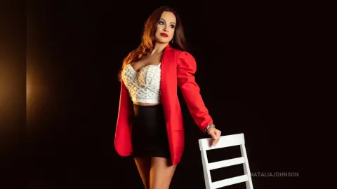 Elegant woman in a red blazer and white lace corset posing confidently against a dark studio background.