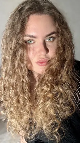 Close-up portrait of a woman with long curly blonde hair and natural makeup in soft sunlight.