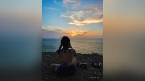 Woman in blue bikini sitting on a cliff overlooking the ocean during a peaceful golden hour sunset.