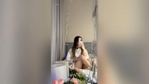 Young woman in a white shirt taking a mirror selfie in a cozy, dimly lit bedroom with fairy lights.