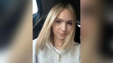 Blonde woman in a grey hoodie sitting in a car with a calm expression.