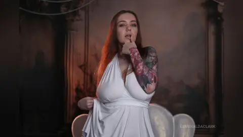EsmeraldaClark poses in a white low-cut dress, showing her long red hair and colorful arm tattoos against a dark, moody background.