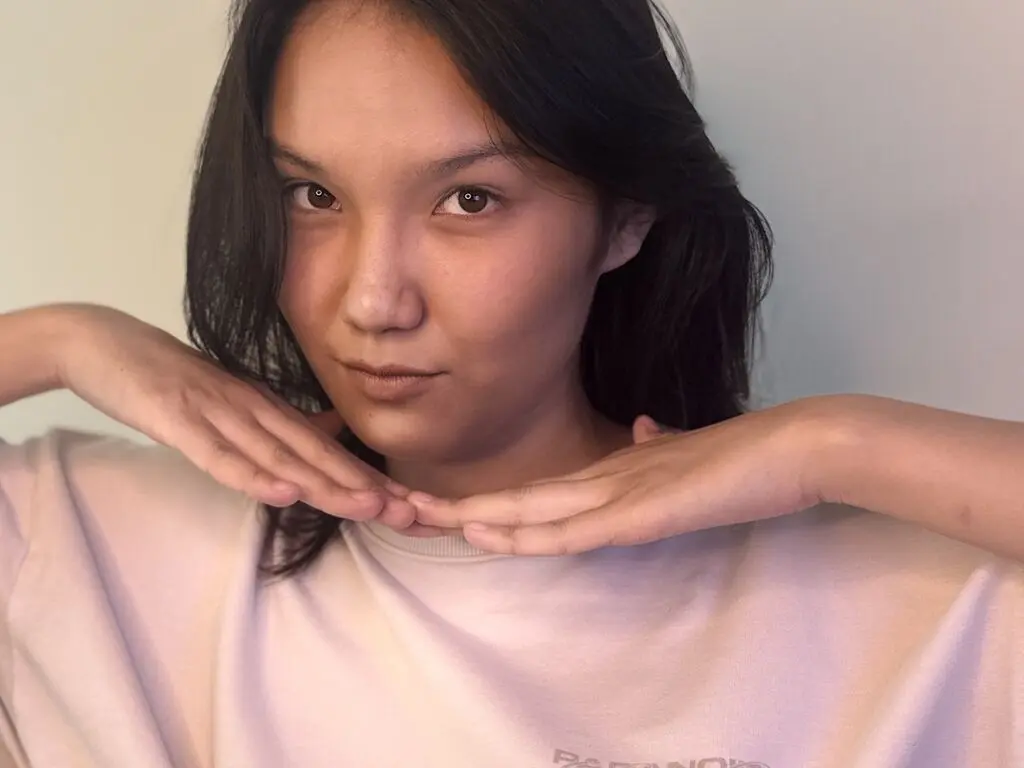 ViolaEbbing poses with her hands under her chin, wearing a light pink sweatshirt and showing her dark hair against a plain wall.