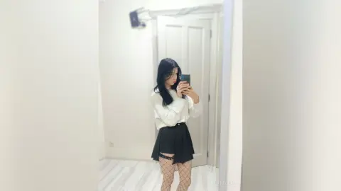 Young woman in a white blouse and black mini skirt taking a mirror selfie in a bright minimalist room.