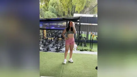 NatiSweet stands outside a gym wearing a grey patterned sports bra and pink shorts with white sneakers.