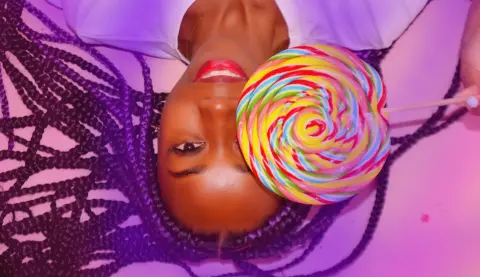 Smiling woman with long dark braids holding a colorful swirl lollipop against a pink background.
