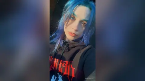 Young woman with blue hair and dark makeup wearing a black graphic hoodie in moody lighting.
