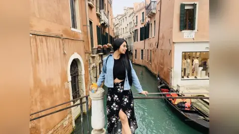 LucyMartin stands on a stone bridge overlooking a narrow Venice canal, wearing a black crop top and a black floral skirt.