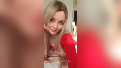DianeDavies poses with long blonde hair, wearing a red long-sleeve top and a heart necklace, looking at the camera.