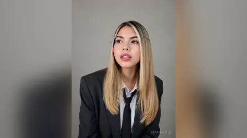 Young woman in a professional black blazer and tie looking thoughtful against a neutral grey background.