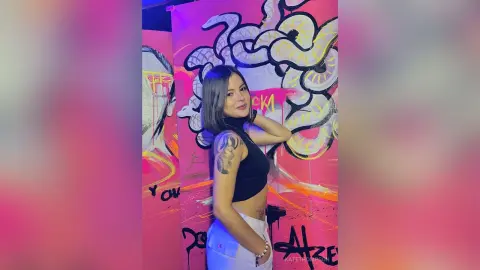 Young woman in a black sleeveless top posing against vibrant pink and white graffiti street art.