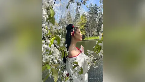 IngridSaint poses in a white dress amidst blooming white flowers by a lake.