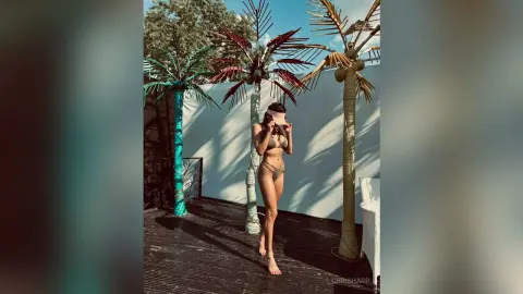 Young woman in a tan bikini taking a selfie by palm trees on a sunny tropical patio.