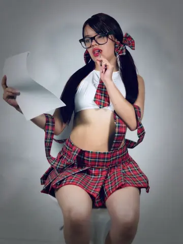 Young woman in red plaid schoolgirl cosplay outfit with glasses holding paper in a studio setting.