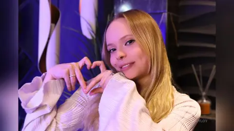 LucyLoy is a blonde woman with a nose ring smiling while making a heart shape with her hands.