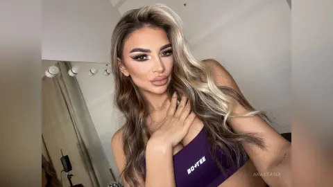 Anastasia poses with her hand on her chest, wearing a purple Bo-Tee sports bra with long, wavy blonde-highlighted hair.