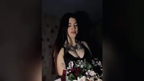 MitsueEastes has long dark hair and a nose ring, wearing a black top that shows off her chest tattoos and cleavage while holding white flowers.