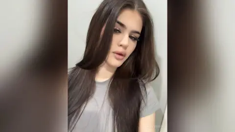 Young woman with long brown hair wearing a grey t-shirt in a soft, minimalist indoor setting.