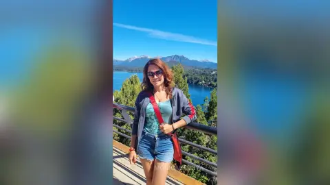 Smiling woman in sunglasses and denim shorts enjoying a sunny mountain lake view from a wooden balcony.