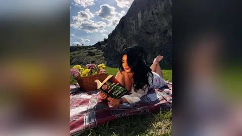 Young woman in a white dress reading a book on a picnic blanket near a mountain under a sunny sky.