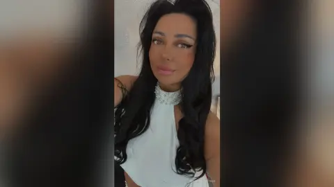 Glamorous woman with long dark hair wearing a white halter top and sparkling rhinestone choker in a bright room.