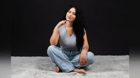 KaiaSinclair sits cross-legged on a white rug wearing a grey v-neck shirt and blue jeans.