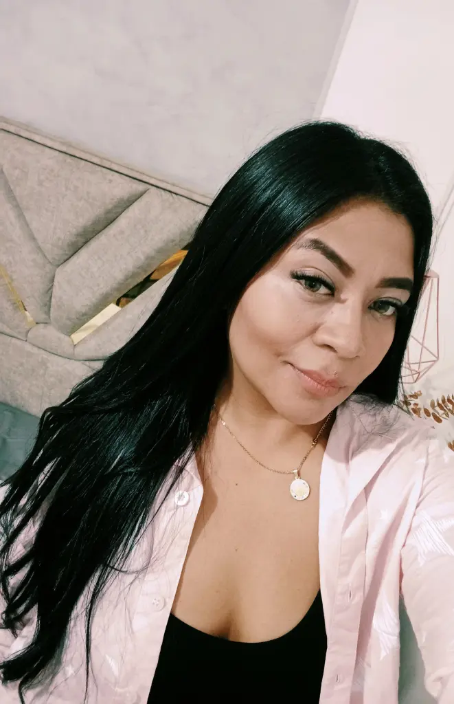 Smiling woman in a sleek black long-sleeve outfit relaxing on a bed in a cozy, dimly lit bedroom.