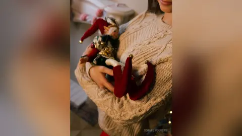 Woman in a cozy cream cable-knit sweater holding a festive holiday elf doll in a warm indoor setting.