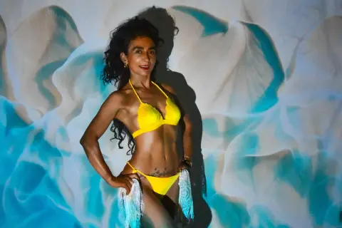 Smiling woman in a bright yellow bikini posing against a dreamy blue and white wavy fabric backdrop.