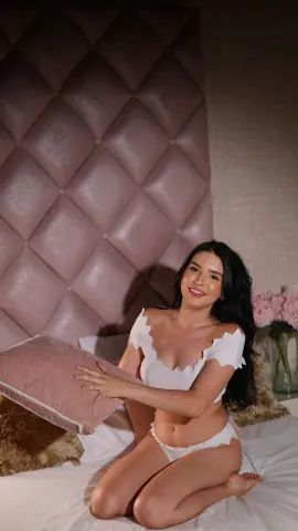 Smiling young woman in white lace lingerie relaxing on a bed with a pink tufted headboard.