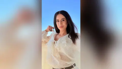 BellaLeen poses with long dark hair, wearing a white polka-dot blouse with a tie-front detail against a clear blue sky.