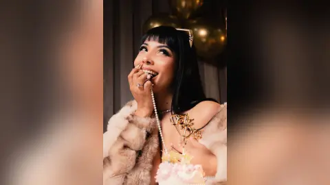 Smiling woman with dark hair in a plush cream faux fur coat wearing gold jewelry at a festive celebration.