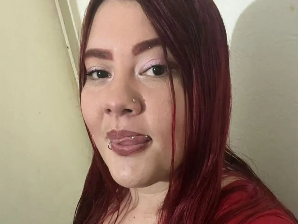 Close-up portrait of a smiling woman with long red hair and nose piercing in a bright indoor setting.