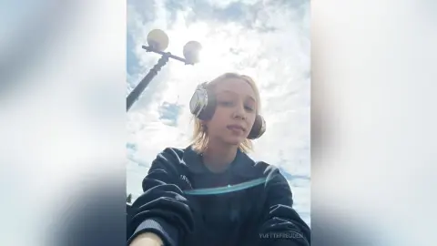Blonde woman in a dark sweatshirt wearing headphones outdoors under a bright cloudy sky.
