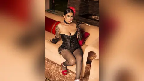 JuliahLewis wears a black lace corset and fishnet stockings while sitting on a tan sofa, holding a cigarette in her mouth.