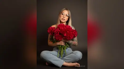 Blonde woman in glasses and denim holding a large bouquet of red peonies against a dark studio background.