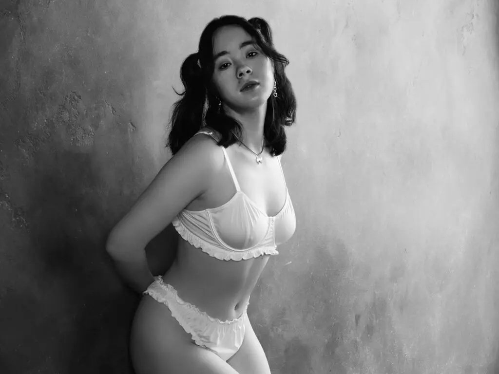 Moody black and white portrait of a young woman in white lace lingerie against a textured wall.