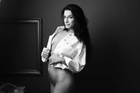 Smiling pregnant woman in an open white shirt posing against a dark background in a moody studio setting.