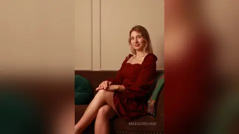 Smiling young woman in a long-sleeved burgundy dress sitting on a sofa in a cozy indoor setting.