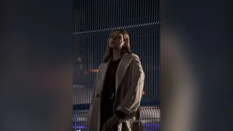 Moody portrait of a woman in a beige coat standing against a metal fence at night.