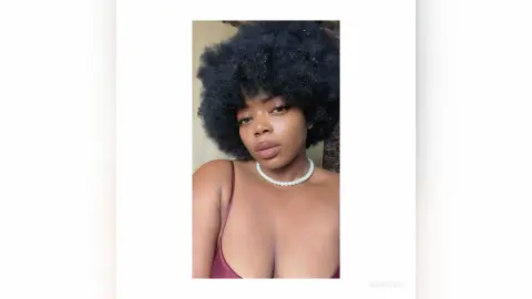 Young woman with voluminous afro hair wearing a pearl necklace and burgundy top in a soft, natural light setting.