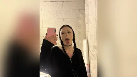 Young woman with dark braids and black zip-up hoodie taking a playful mirror selfie in a minimalist room.