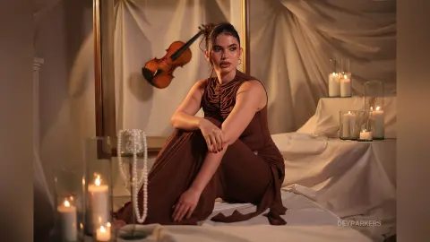 Elegant woman in a brown dress sitting on a bed surrounded by glowing candles in a warm, moody room.