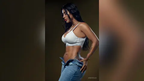 AnnyRoug poses in a white Calvin Klein bra and unbuttoned blue jeans, showing her toned stomach and hip tattoo against a dark background.